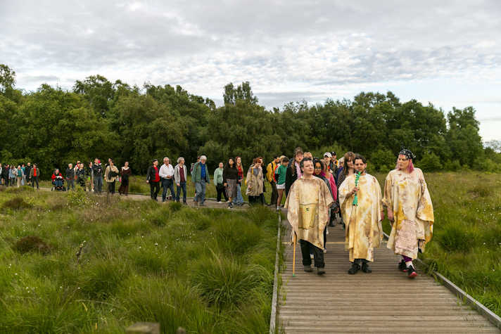Three people wearing off white cloaks lead a group of people through a boggy peatland on a wooden walkway.