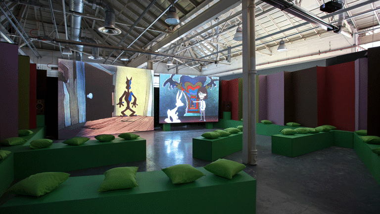 Installation view of an exhibition showing two projection screens with films, and rows of green benches with green cushions on