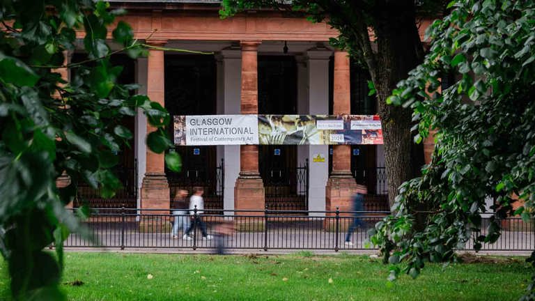 Sandstone, columned building viewed through leafy trees and grass, with a Glasgow International festival banner hanging between the columns and blurred figures walking past.