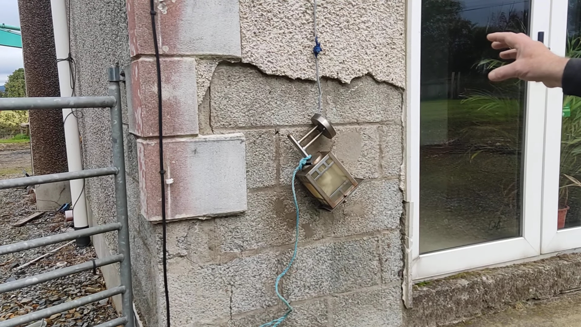 A hand points towards the exterior wall of a domestic dwelling, showing exposed brickwork. A small metal device hands on the wall, suspended by blue string.