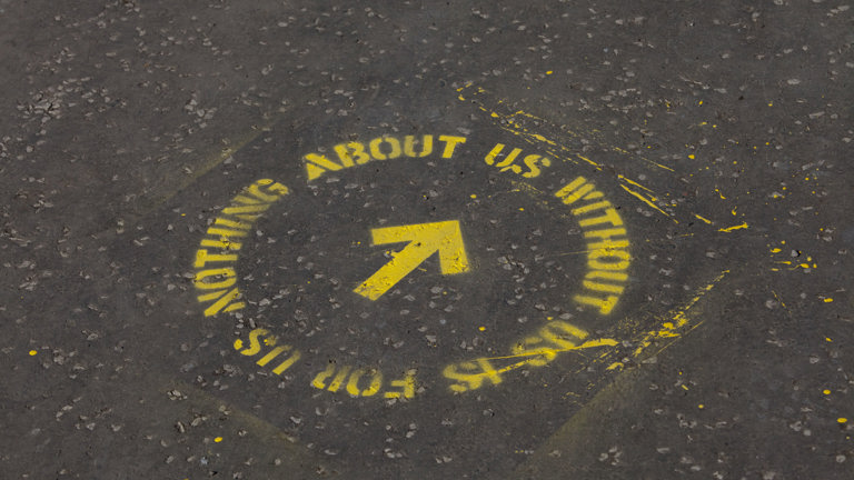 yellow spray paint stencilled onto asphalt floor. it reads 'nothing about us without us is for us' in a circle with an arrow in the middle