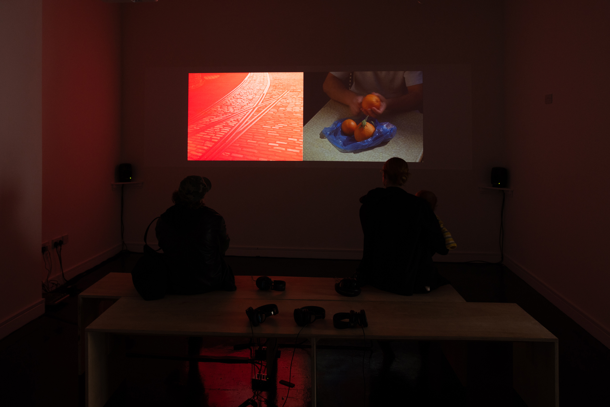 Two visitors seated on a low platform, silhouetted against a split-screen video projection showing red-toned cobbled tram tracks alongside footage of hands peeling oranges, with headphones laid out on the bench in front of them.