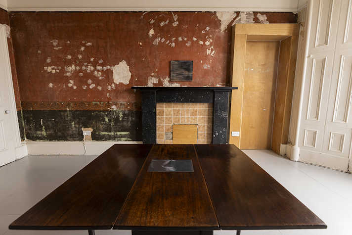 A photo is displayed above a fireplace in a room, which has peeling red wallpaper. In the centre of the room is a wooden table with a painting displayed.
