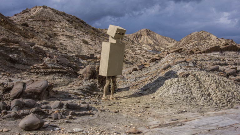 Film still showing a rocky landscape and a figure in box-shaped costume