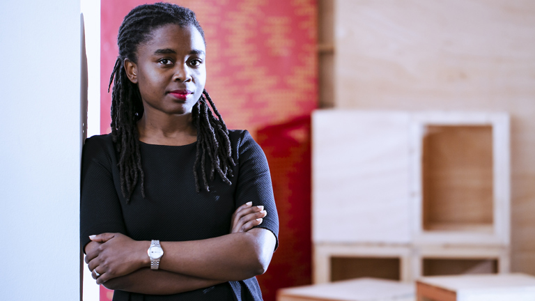 A woman with braids, wearing a black top, leans against a doorway with her arms crossed.