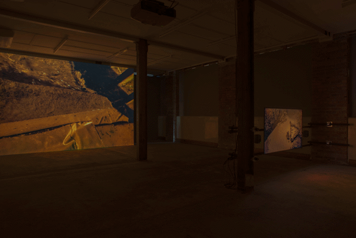 a dark industrial space with two films projected. On the screens are abstract images of earth.