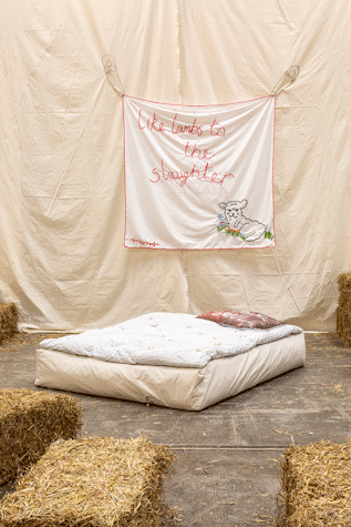 A stuffed mattress is in front of artwork hanging on a wall of fabric. Surrounding the mattress are straw bales.