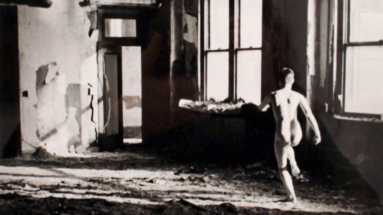  A black and white photograph of a naked person running  across a room, away from the camera, towards a partially open  door. They are in an abandoned building with debris on the floor  and paint peeling from the walls.