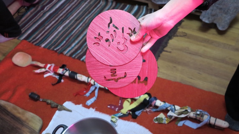 A hand lit by red light holds three circular laser-cut wooden discs. In the background, a long wooden spoon and various cooking utensils rest on white, red, and multicoloured rugs spread across the floor. 