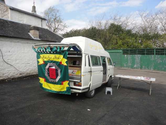 parked white camper van with a square banner hanging from the lifted boot door which reads aye aye books