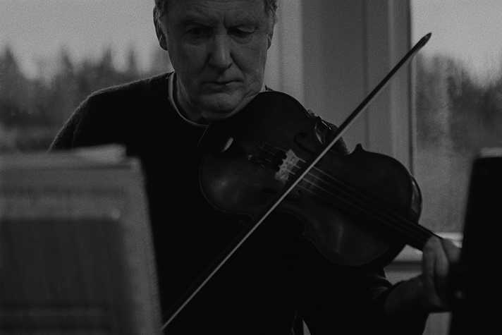 B+W close up image of a man in his 50s with sandy hair playing violin and looking intently at the music.]