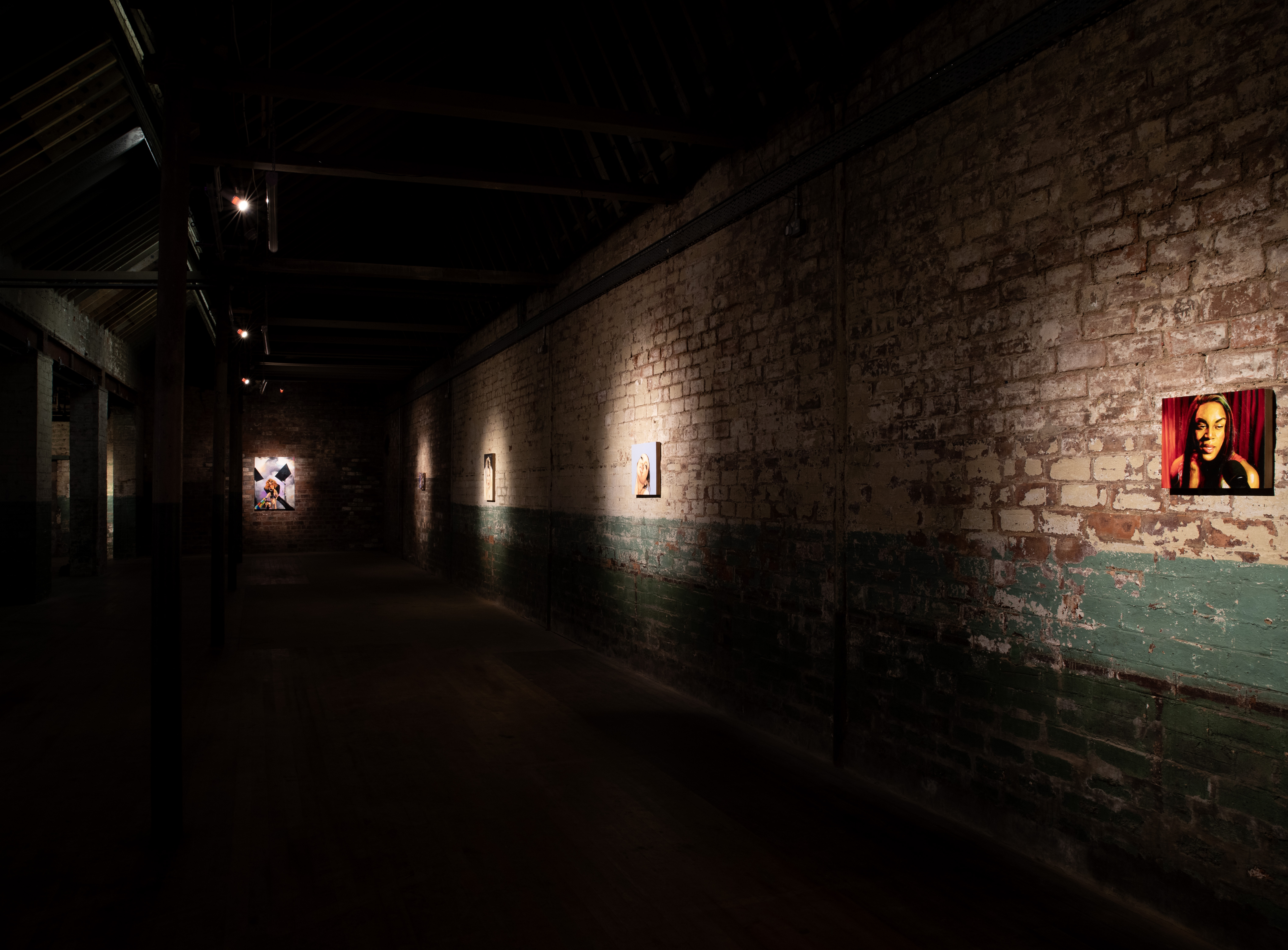 Long dimly lit gallery corridor with exposed brick and green-painted lower walls receding into the distance. A row of small spotlit portrait paintings of women runs along the right-hand wall, with a larger canvas visible further down the corridor on the left.