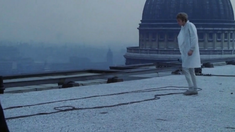 A film still showing a figure on a roof top with a large dome behind 