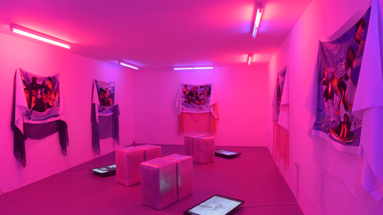  A room with pink neon lights has three screens on the floor, next to sitting blocks and on the walls it has five fabric hanging with images of women on it. 