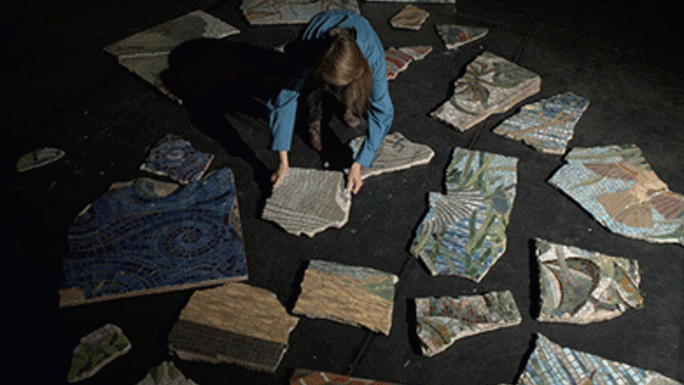 Film still, person kneeling on floor arranging pieces ofceramic artwork