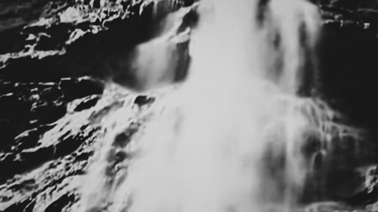 A black and white still featuring water pouring in front of a textured rocky surface