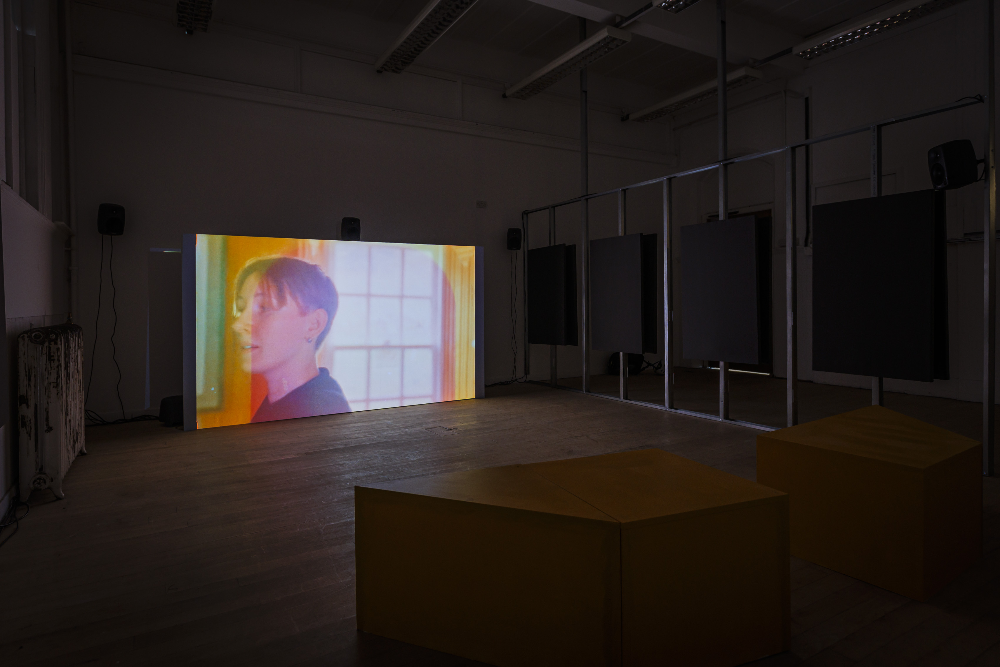 In a blacked out room, a glowing projection screen is visible. The rest of the room's architecture is not visible, apart from the illuminated edges of a metal frame wall bearing sound panels. On the screen, a still image is projected of a person's head looking off screen to the left. Behind the person a window is visible with light streaming in.  