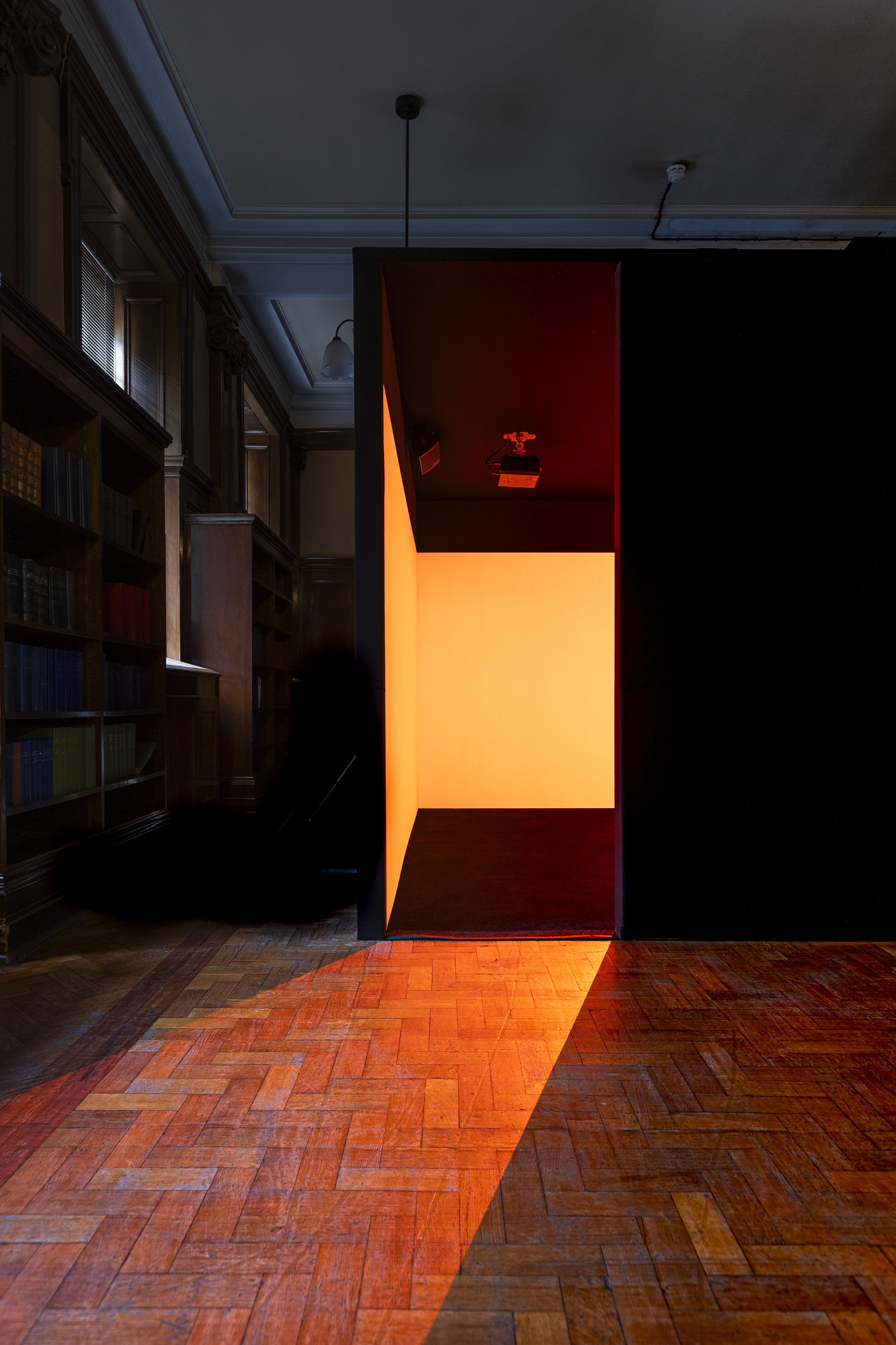 A dark box-like structure glowing orange from within stands in the Mitchell Library's reading room, its light spilling across the herringbone parquet floor, part of Irienu Destourelles' installation.
