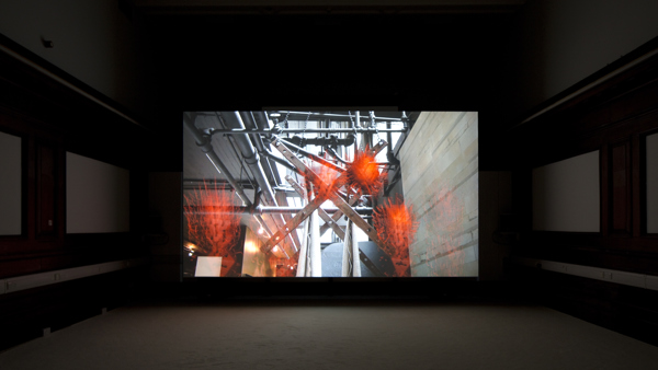 In a dark room, there is a large projector screen placed in the centre. This screen shows the image of a wall on the side and pipes spread out throughout the area. There are some orange translucent work like objects, made of hay like material, placed on various parts of the image. The walls on the sides of the screen have some screens hanging on them, but they are barely visible due to the darkness.