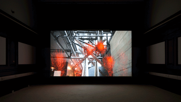 In a dark room, there is a large projector screen placed in the centre. This screen shows the image of a wall on the side and pipes spread out throughout the area. There are some orange translucent work like objects, made of hay like material, placed on various parts of the image. The walls on the sides of the screen have some screens hanging on them, but they are barely visible due to the darkness.