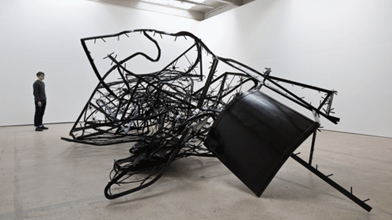 A white gallery with a large tangled metal sculpture. A figure is standing to the left 