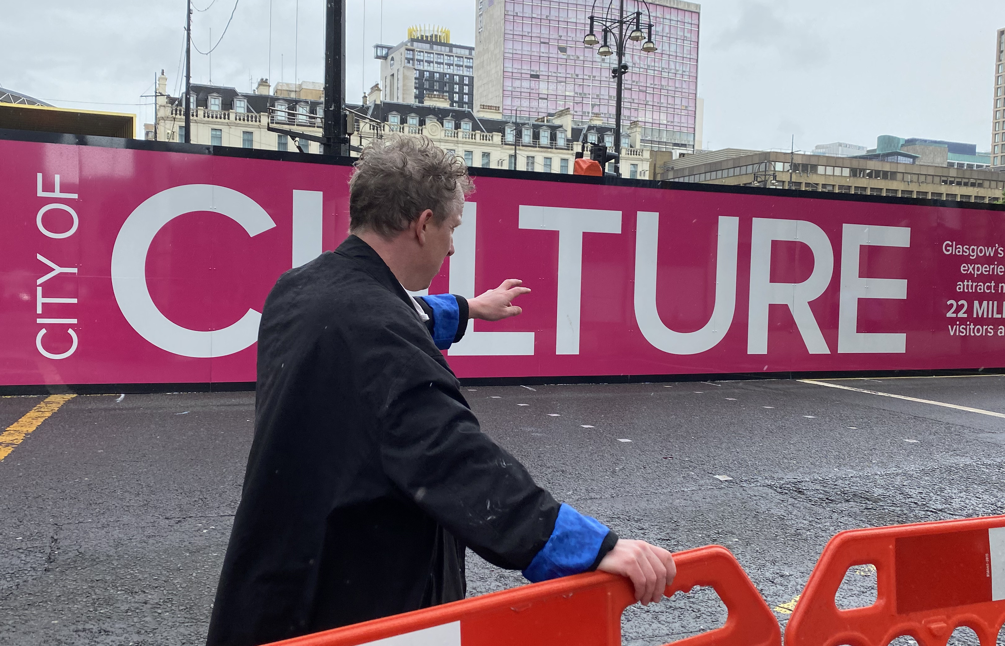 Colour photograph of a man with his back to the camera gesutring toward the George Sqaure construction site. In the background, a pink banner reads "City of Culture," and he holds an orange traffic barrier. 