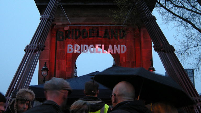 group of people stood on a bridge. on the bridge there is a sign that says bridgeland.