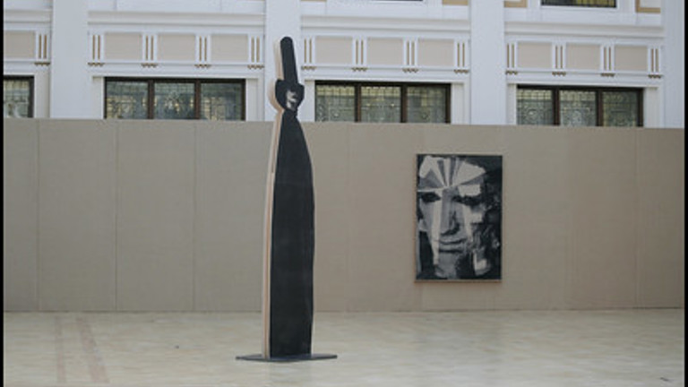 installation featuring a life-sized cutout panel with black-and-white photographic image of a cloaked figure. behind is an abstract painting. in a bright and airy atrium of he Mitchell library.