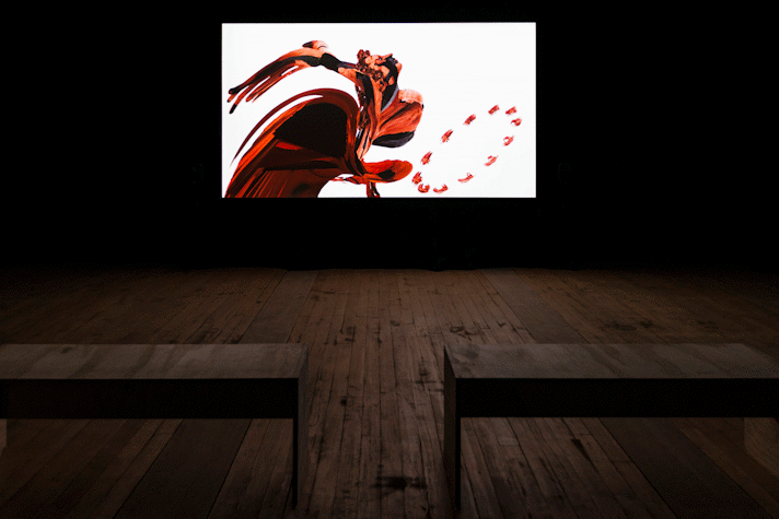 Film is projected onto a screen in a dark room. There are wooden benches facing the screen.