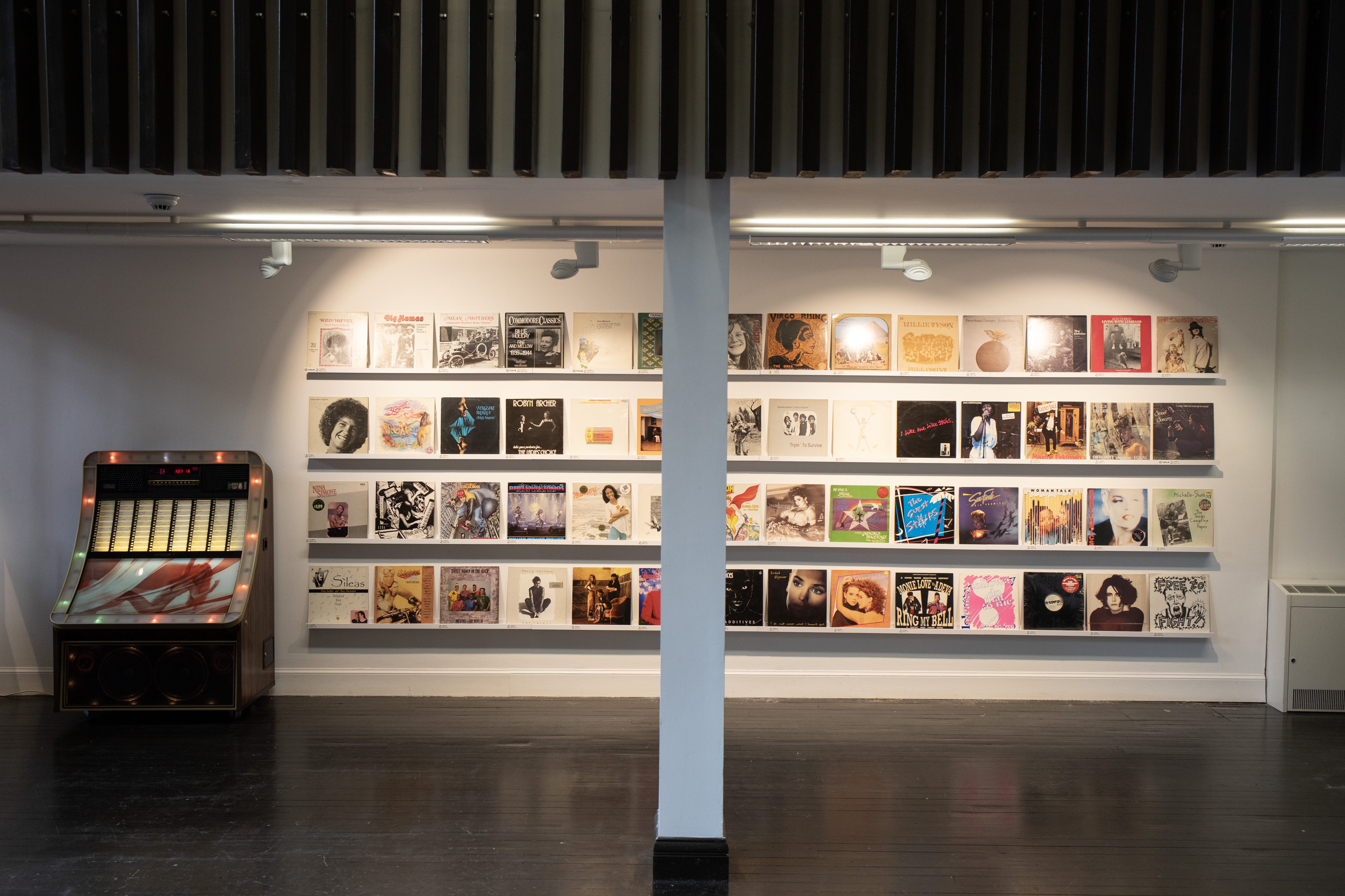 A gallery wall displaying rows of vintage record sleeves by women musicians, with an illuminated jukebox standing beside them on a dark wooden floor.