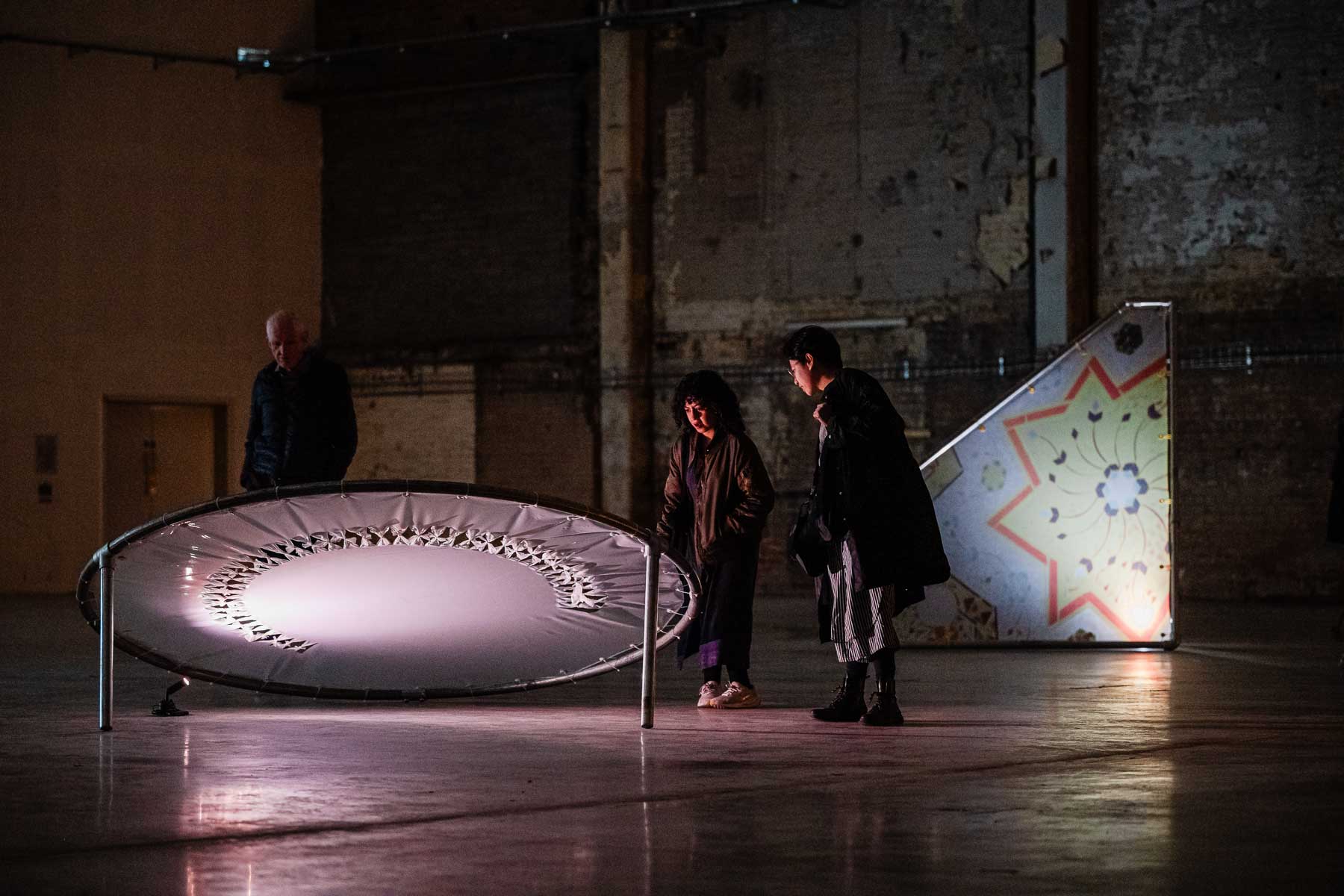 Visitors gather around illuminated sculptural installations in a dim industrial gallery, examining the artworks in a quiet exhibition space.