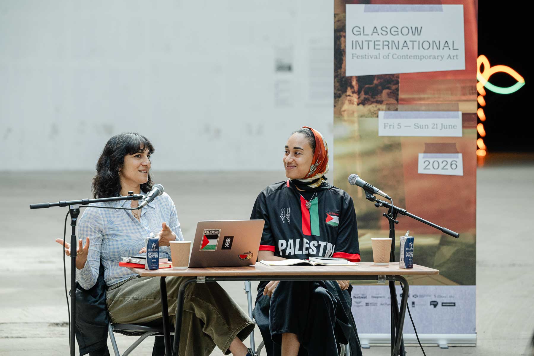 Two women sit at a small outdoor table with a laptop, one speaking with her hands raised, the other wearing a headscarf and a "Palestine" football shirt. A Glasgow International festival banner and neon sign are visible behind them.