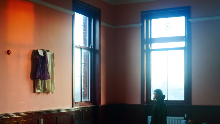 Colour photograph of a woman standing in a large room with peach-coloured walls. Two small fabric pieces hand on the wall to the left. Large windows cast a blue hue across the space. 
