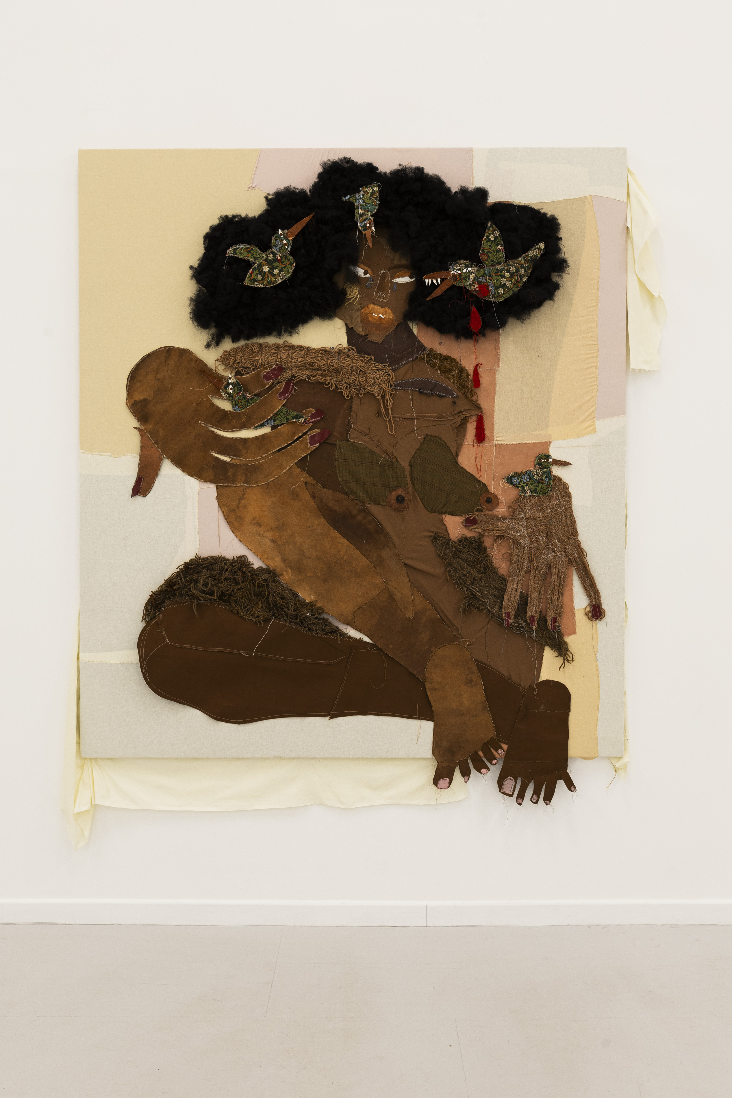 A large mixed-media textile portrait of a seated figure with voluptuous woollen black hair, patterned fabric birds with sharp beaks nesting in the hair, and oversized stitched hands and feet against a patchwork beige and pink ground.