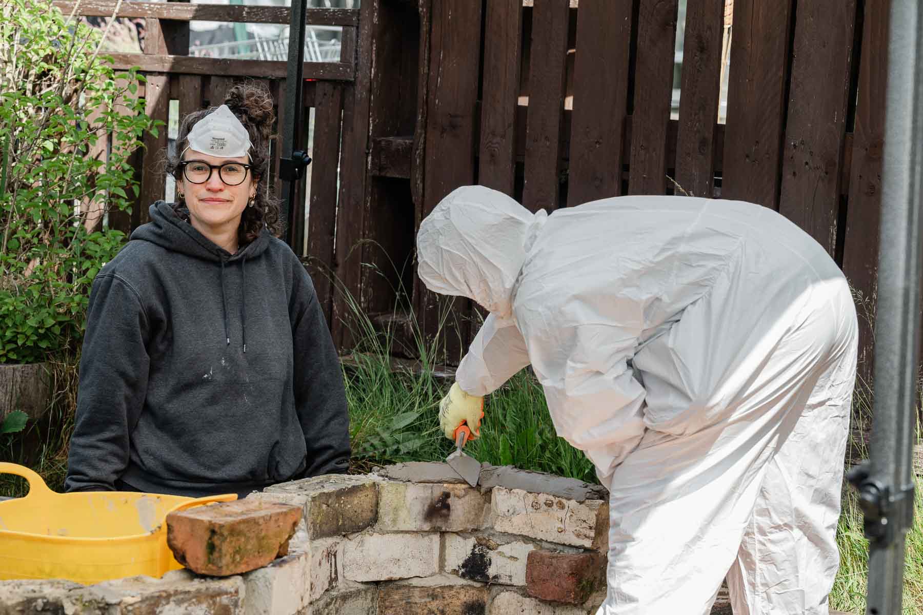A woman in a grey hoodie and dust mask smiles at the camera while another person in a white protective suit works with a trowel on a low brick wall in a garden.