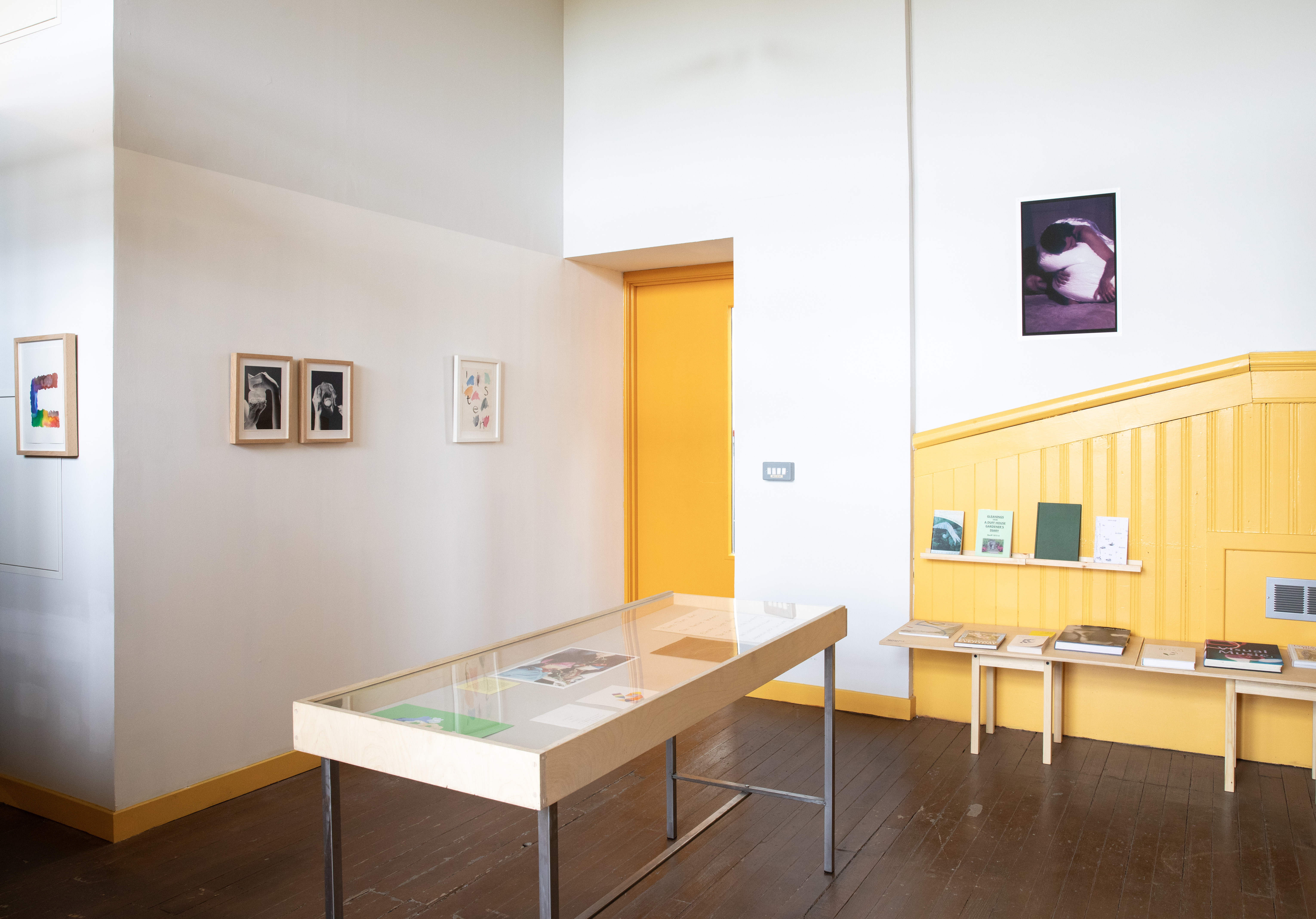 Wide gallery view with white walls meeting a yellow-panelled corner and doorway. Framed works line the left wall, including a colourful drip painting, two black-and-white photographs, and the "listen" piece, with a photograph of a curled-up figure on the right wall. A glass vitrine holds printed materials, and a yellow shelf displays zines and books along the panelled wall.