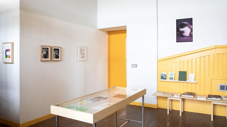 Wide gallery view with white walls meeting a yellow-panelled corner and doorway. Framed works line the left wall, including a colourful drip painting, two black-and-white photographs, and the "listen" piece, with a photograph of a curled-up figure on the right wall. A glass vitrine holds printed materials, and a yellow shelf displays zines and books along the panelled wall.
