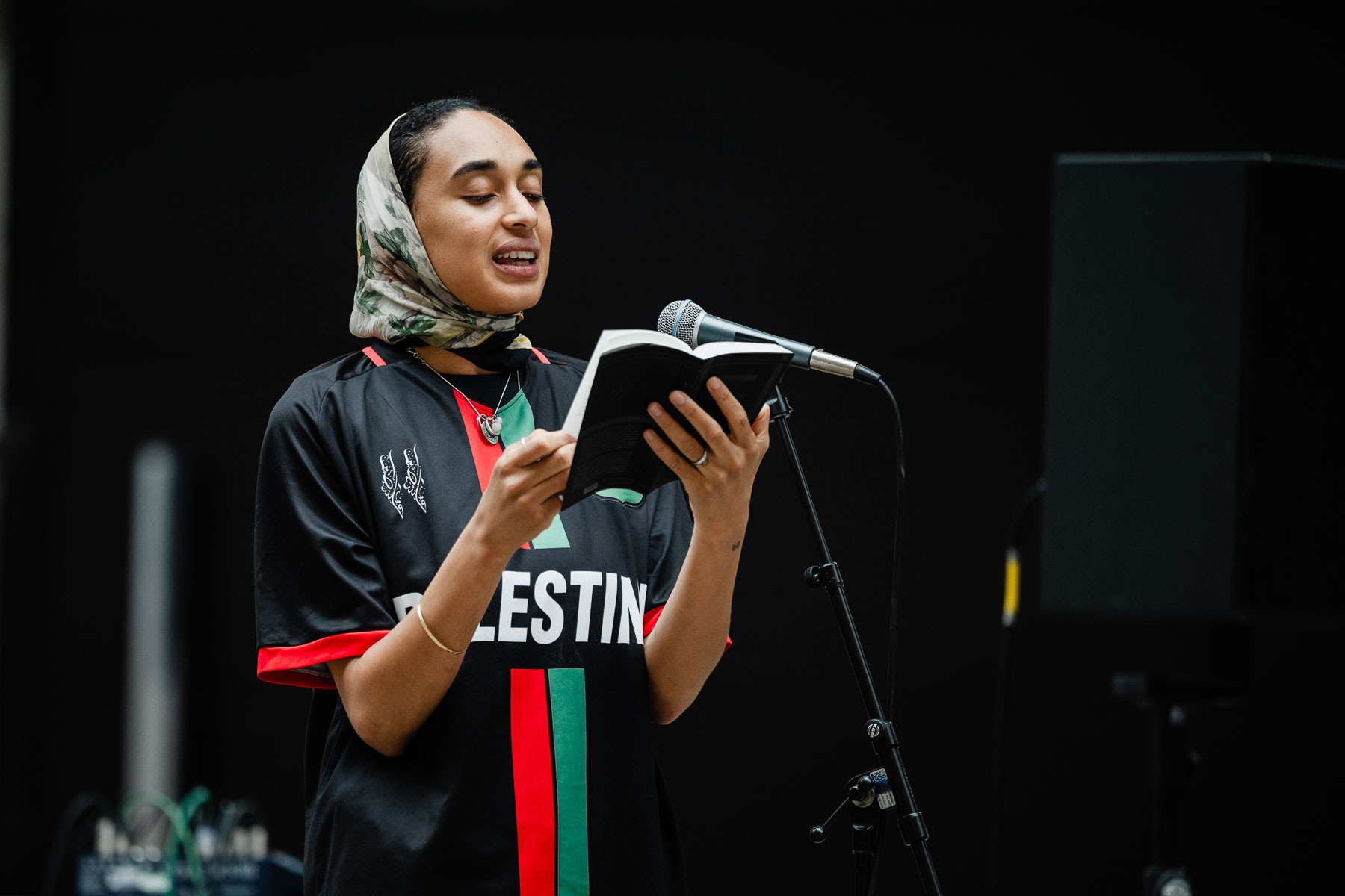 Close-up of a woman wearing a headscarf and a black, red and green "Palestine" football shirt, reading aloud from a book into a microphone against a dark background.