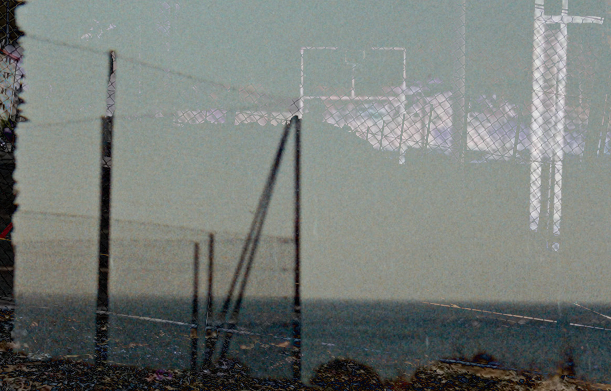  A grainy analogue image of a fence in the foreground with a distant blue-tinged horizon behind it. Highlights of a second image are overlaid, though the details are indistincy. The diamon pattern of the fencing dominates the composition. 