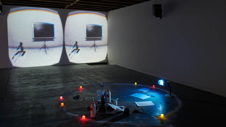 An installation view of a gallery with projections on the back wall, with coloured lights and objects on the floor in the foreground