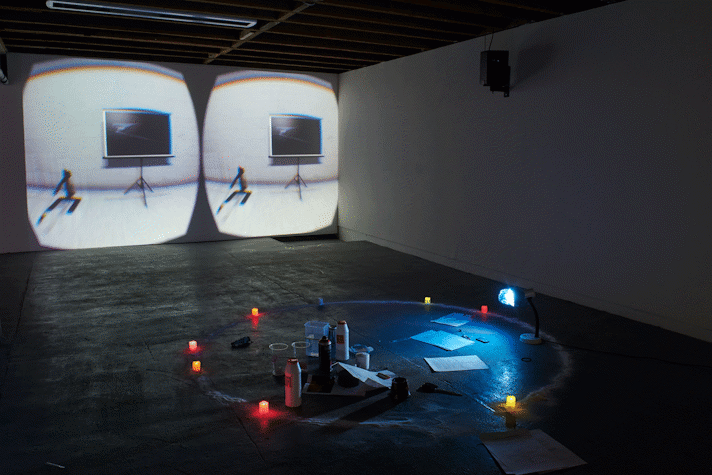 An installation view of a gallery with projections on the back wall, with coloured lights and objects on the floor in the foreground