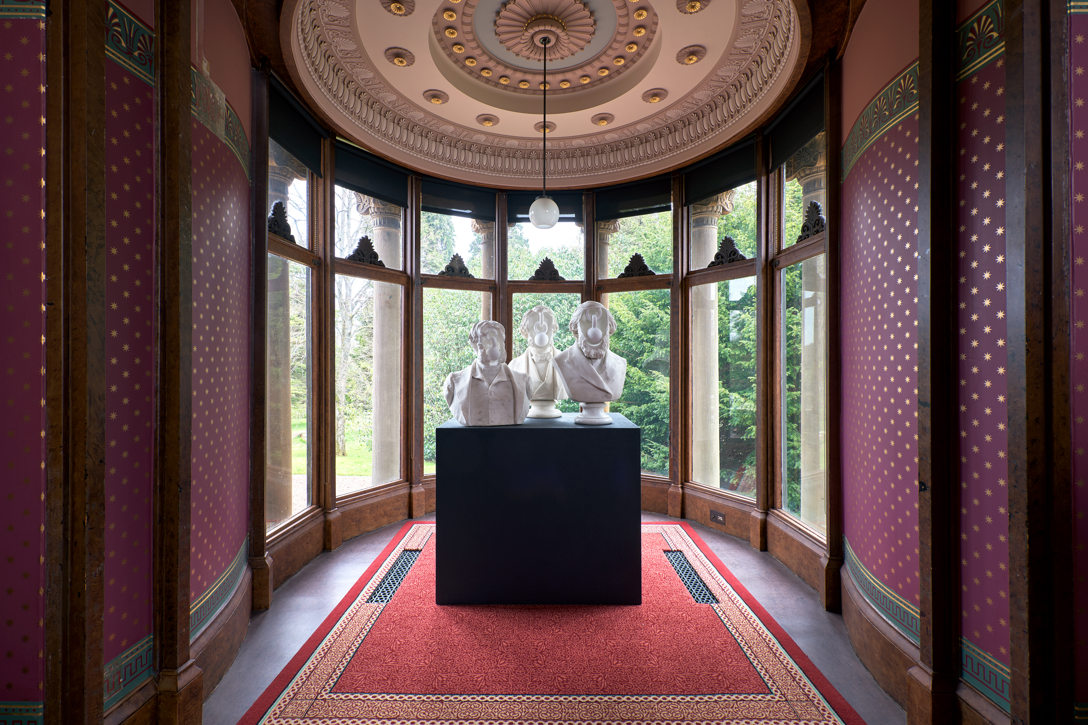 Three white marble busts on a black plinth in Holmwood House's bay window, set against star-patterned wallpaper and an ornate domed ceiling, part of Jon Owen's installation.