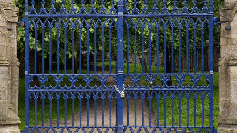 A pair of ornate wrought iron gates painted blue, about 10 feet tall, are pictured closed with a large padlock and chain. Behind the gates a path flanked by grass can be seen, with some stone plaques dotted among the grass marking grave sites.  