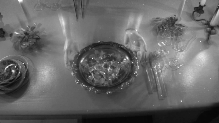 Film still showing an overhead view of a table. Two semi-transparent hands hold a silver dish containing silver-coloured food, surrounded by cutlery, glassware, and candles. 