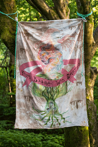 A fabric banner with artwork on it hangs from a tree, it features writing that reads "From Bogge We Emerge"