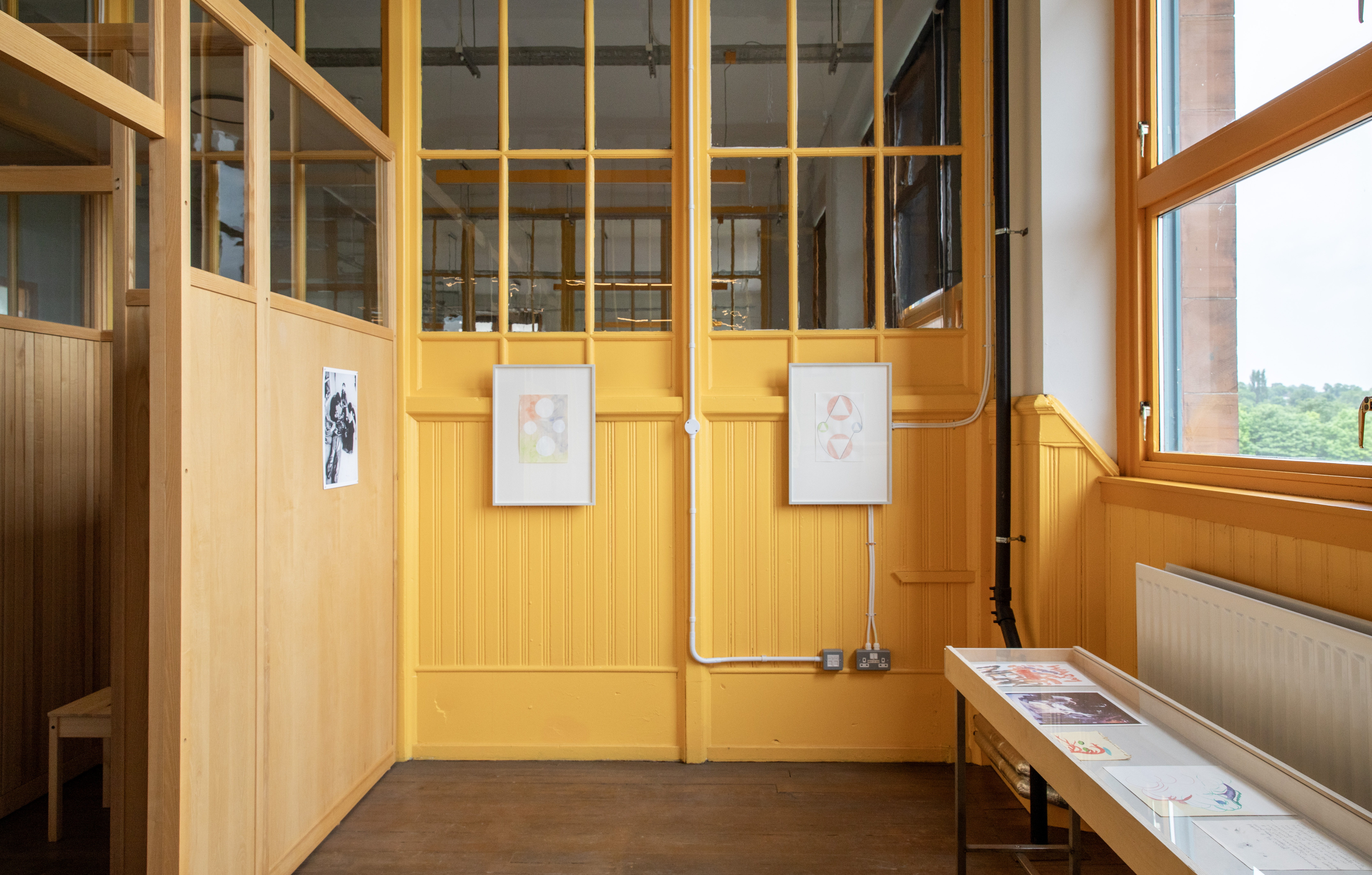 Interior view of a room with yellow-painted wood panelling and glazed doors. Two small framed drawings, one with pale circles, one with geometric triangle shapes, hang side by side. A black-and-white photograph is pinned to a plywood wall on the left, and a table on the right holds several flat printed works.