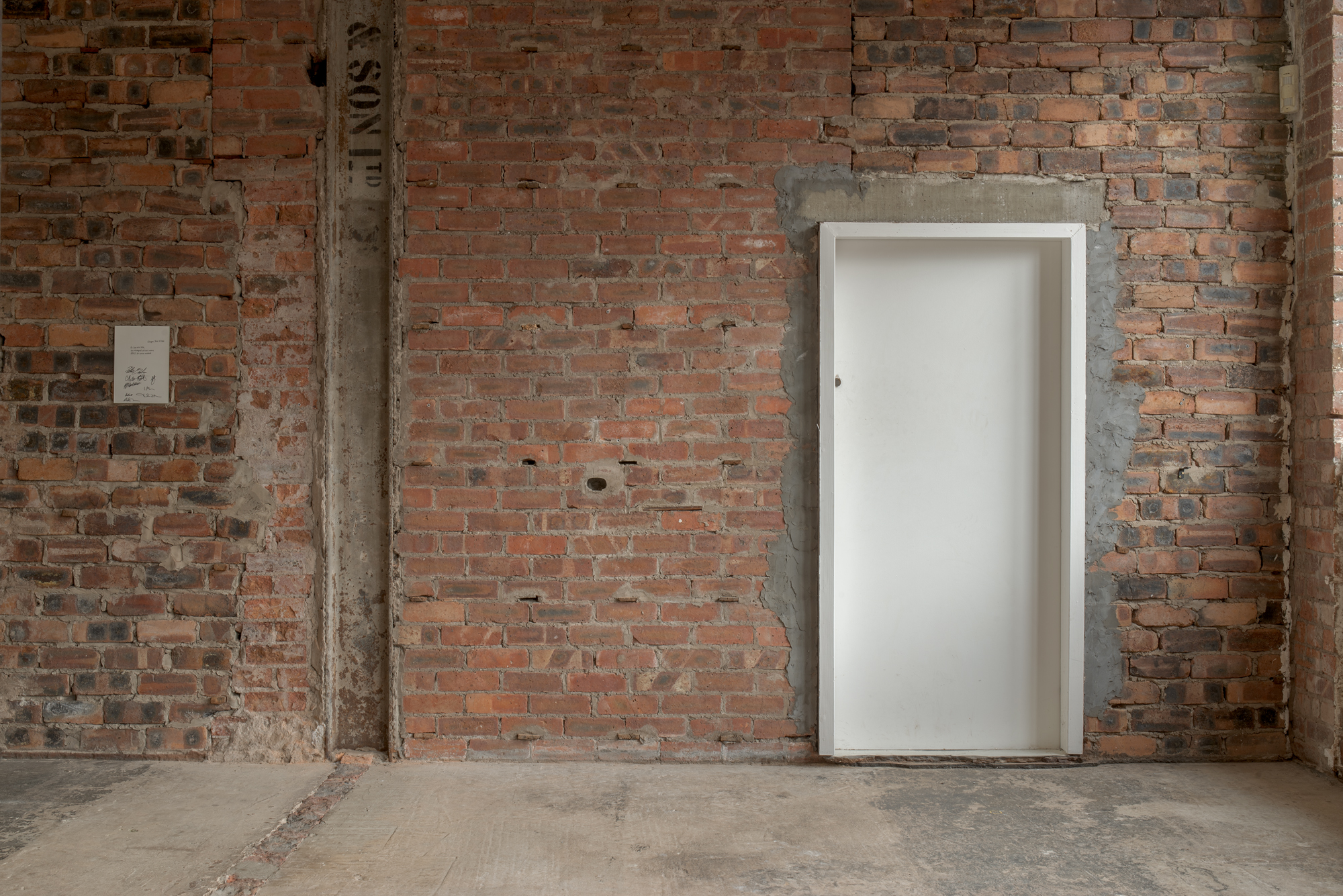 A modern white door set into an exposed brick wall bearing faint stencilled lettering, in a stripped-back industrial interior.