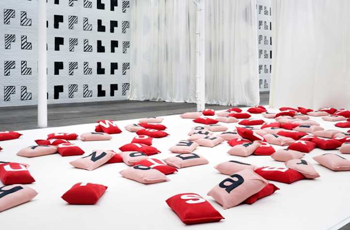 pink and red cushions on the floor. each has a singular letter from the alphabet on it. the cushions lie on top white fabric. in the background there are white curtains and the walls are patterned with L shape blocks.