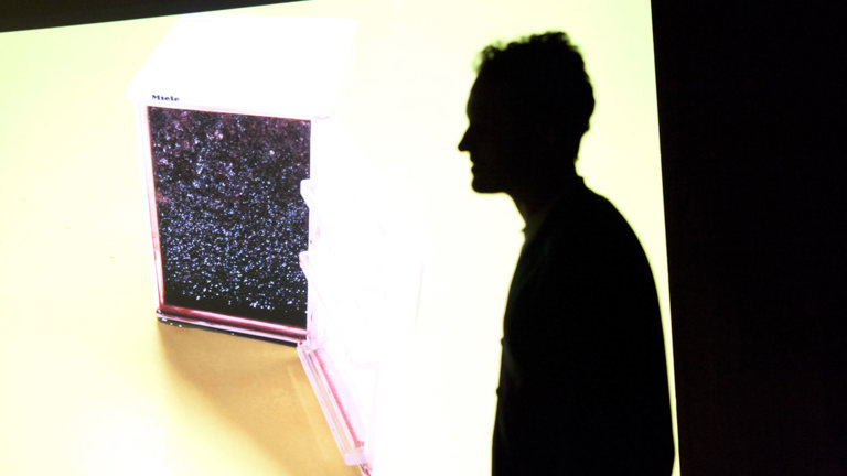 silhouette of a person in front of a large projection. the projection appears to show a small Miele fridge which is open revealing the night sky.