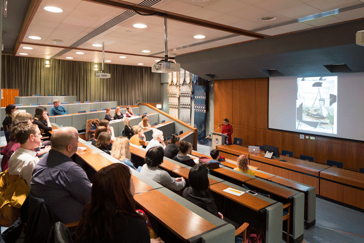 There are multiple people sitting at stationary benches in a lecture theatre. They are facing a projector screen, which is showing an image on it and a woman standing behind a podium with a microphone in front of her.
